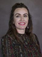 A woman with long brown hair wearing a patterned blouse smiles at the camera against a plain background.