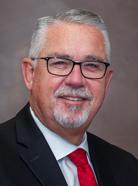 A man with short gray hair and glasses, wearing a black suit, white shirt, and red tie, smiles at the camera against a plain background.